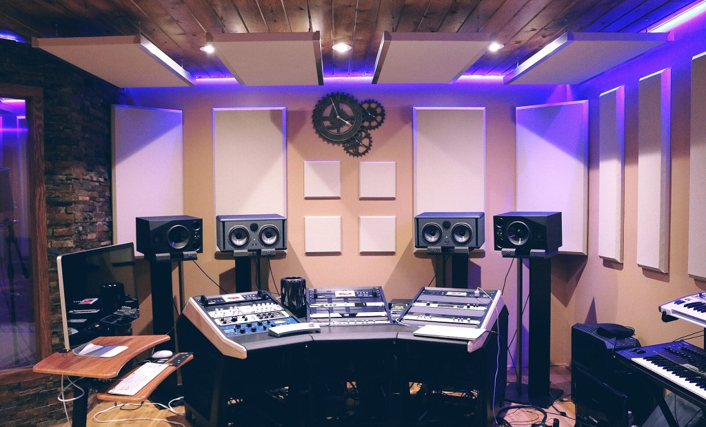 Photo: Dayron Villaverde, CC0, via Wikimedia Commons A modern music studio featuring a curved mixing desk, two studio monitors on stands, a computer, and various audio equipment, illuminated by LED lighting.
