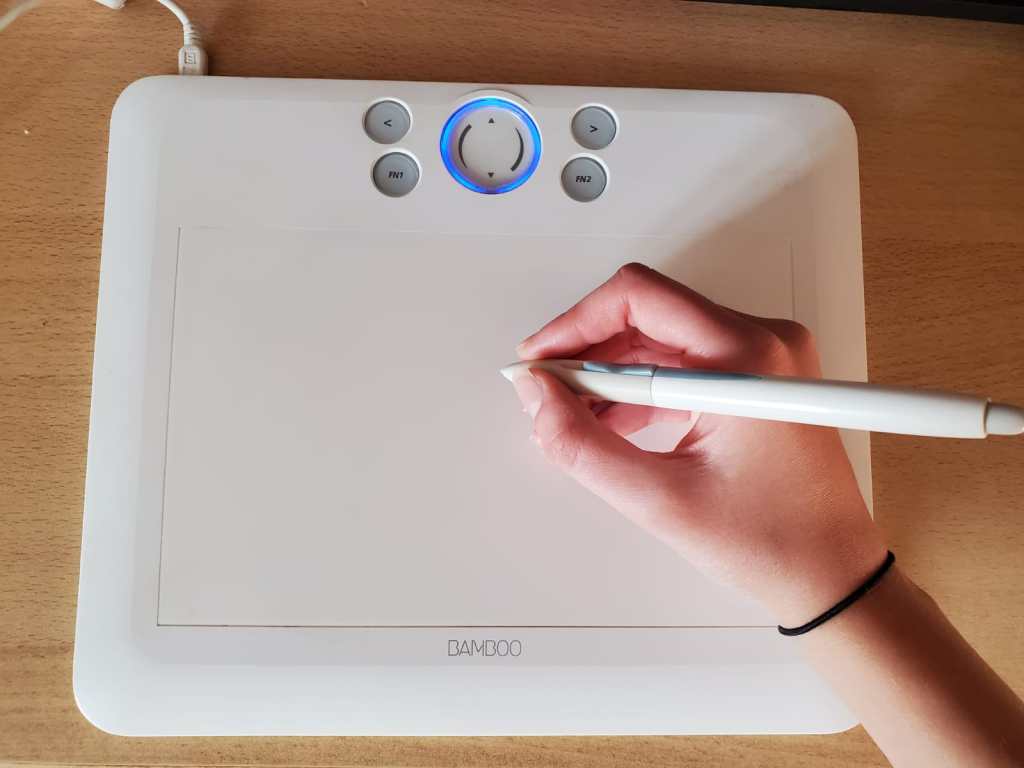 A hand holding a stylus above a white drawing tablet, featuring buttons and a circular touch area, placed on a wooden surface.