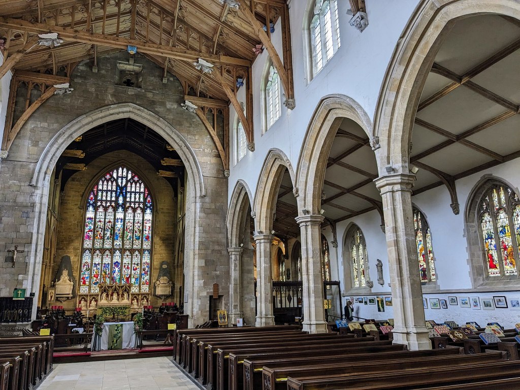 Interior of a church showcasing high wooden ceilings, arched stone columns, and colorful stained glass windows, with wooden pews and religious artifacts visible.
