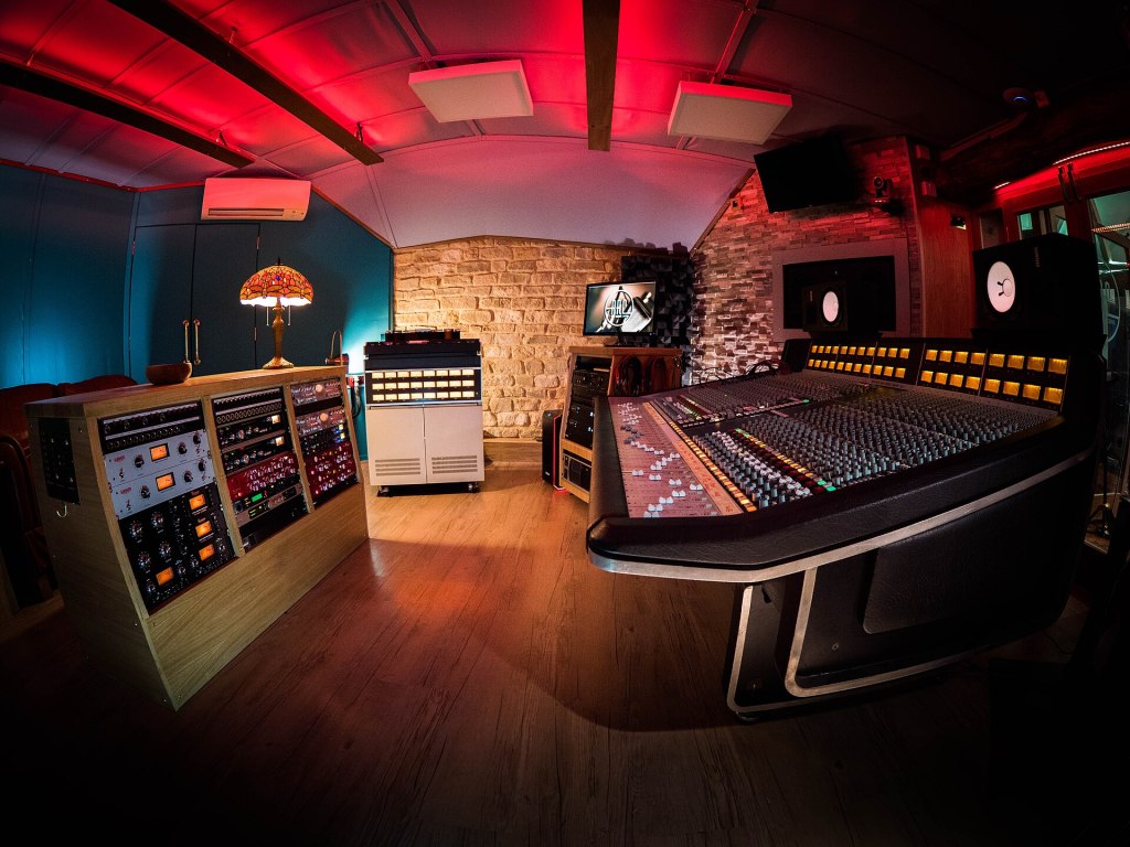 A modern music studio featuring a large mixing console with various controls, surrounded by audio equipment and decorative lighting.