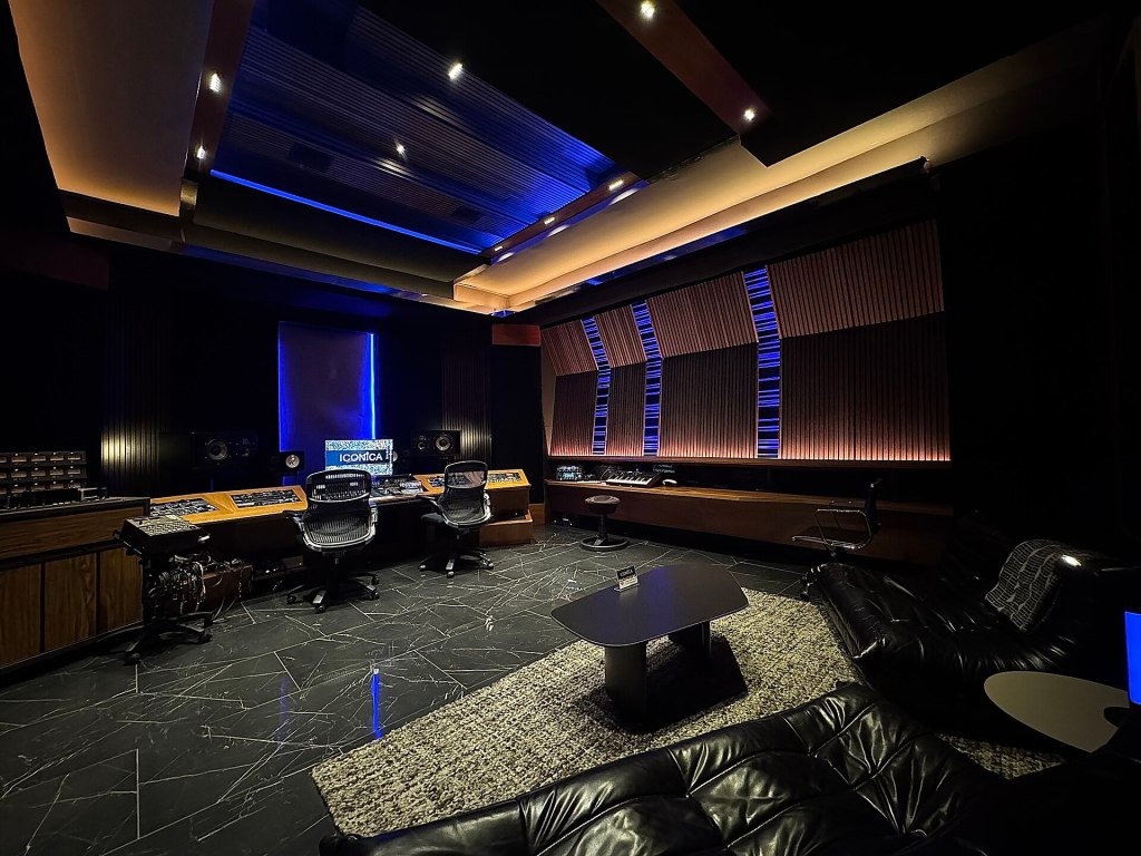 A modern recording studio featuring dark walls, ambient blue lighting, and professional audio equipment. The room includes a mixing console, comfortable seating, and a cozy rug.