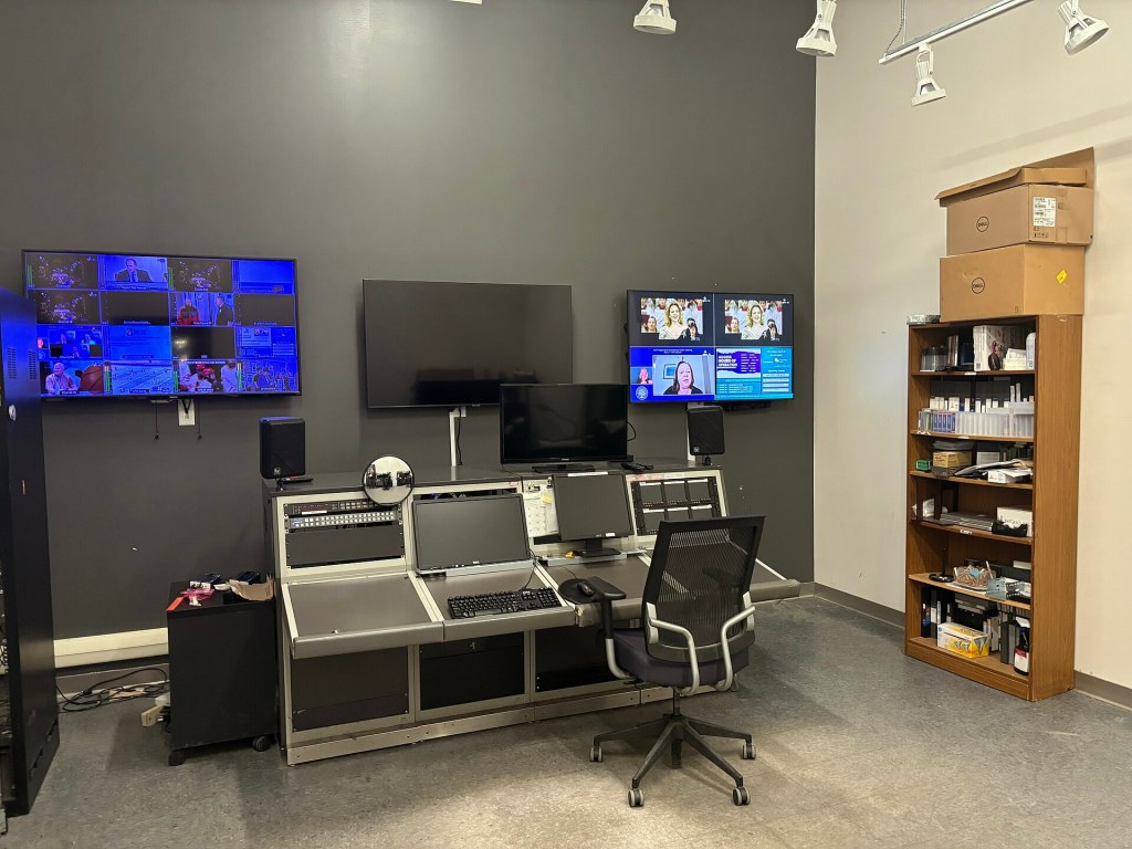 A modern control room featuring multiple monitors displaying different video feeds, a desk with a computer setup, and a chair. To the right, there is a wooden shelf filled with various items.