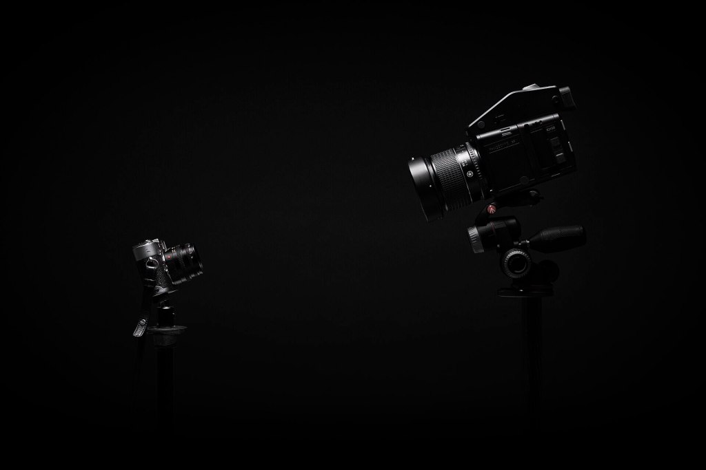 Two cameras on tripods against a black background, one large digital camera on the right and a smaller camera on the left.